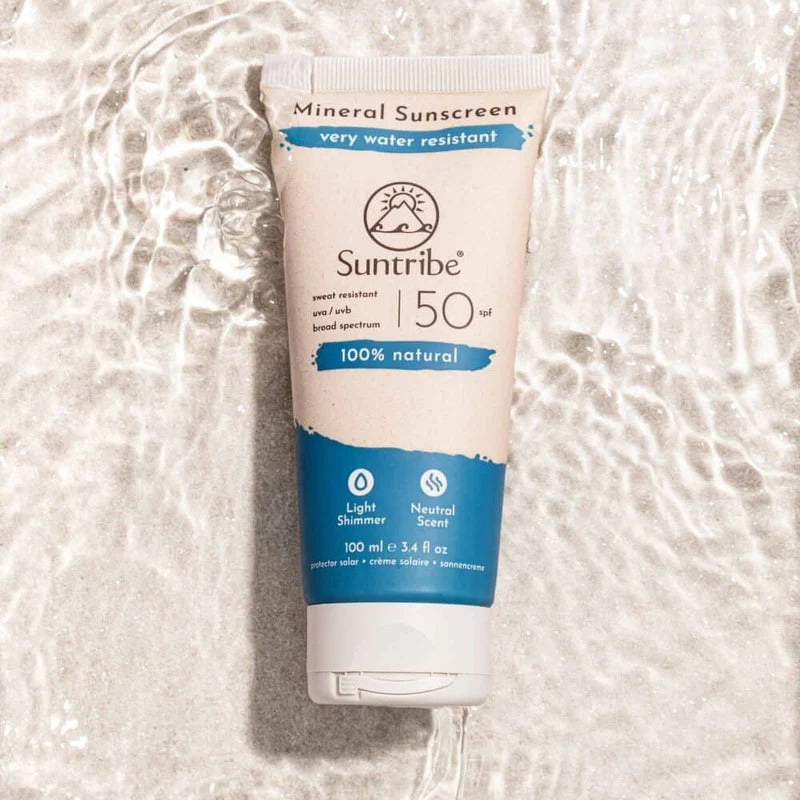 Tube med Suntribe Mineral Sunscreen SPF 50 ligger i grunt vann med sollys som reflekteres. Tuben er beige med blå bunn og hvit kork, og teksten viser at den er 100 % naturlig, vannresistent og parfymefri.