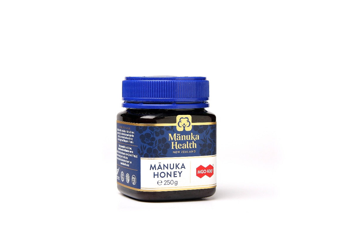 Et mørkt glass med blå skrukork og etikett i blått og gull, merket "Manuka Health New Zealand". Etiketten viser "MGO 400+" og teksten "Manuka Honey 250g", som indikerer høykvalitets manukahonning fra New Zealand.