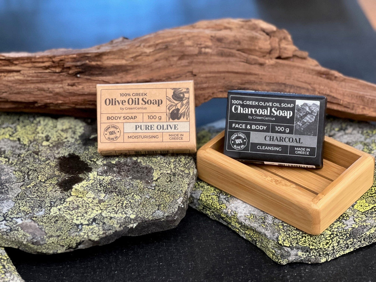 GreenGenius Olivensåpe Kull 100g i papir, sammen med en lys variant på en stein med drivved i bakgrunnen