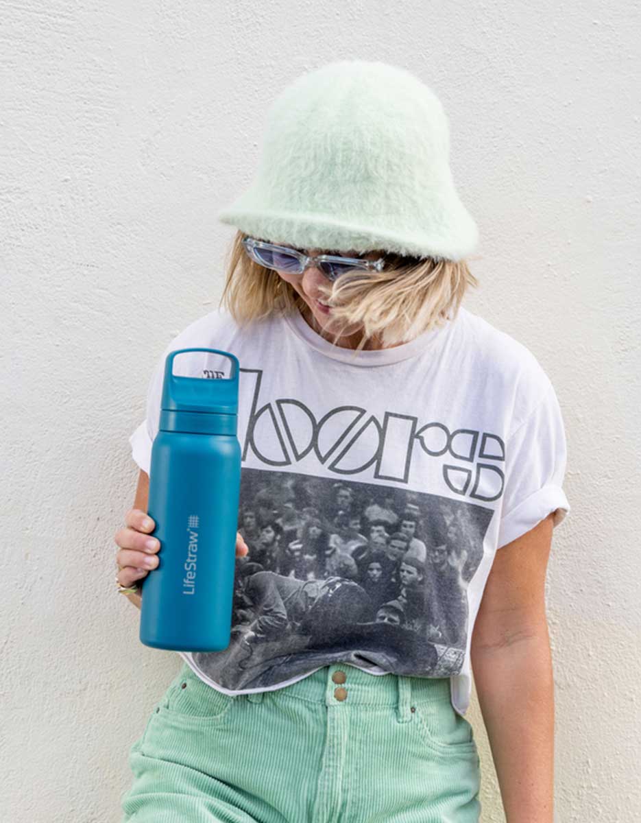 Person med lyst hår, grønn bøttehatt og solbriller holder en blå LifeStraw vannflaske med sugerør foran en lys vegg. Personen har på seg t-skjorte med trykk og lysegrønne bukser.