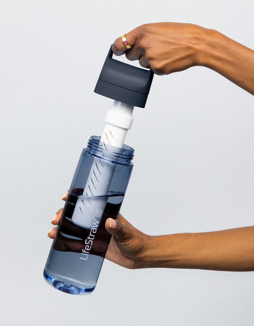 To hender demonstrerer hvordan man setter inn et hvitt vannfilter i en gjennomsiktig blå vannflaske fra merket LifeStraw, mot en lys bakgrunn. Flasken er halvfull med vann og har sort lokk.