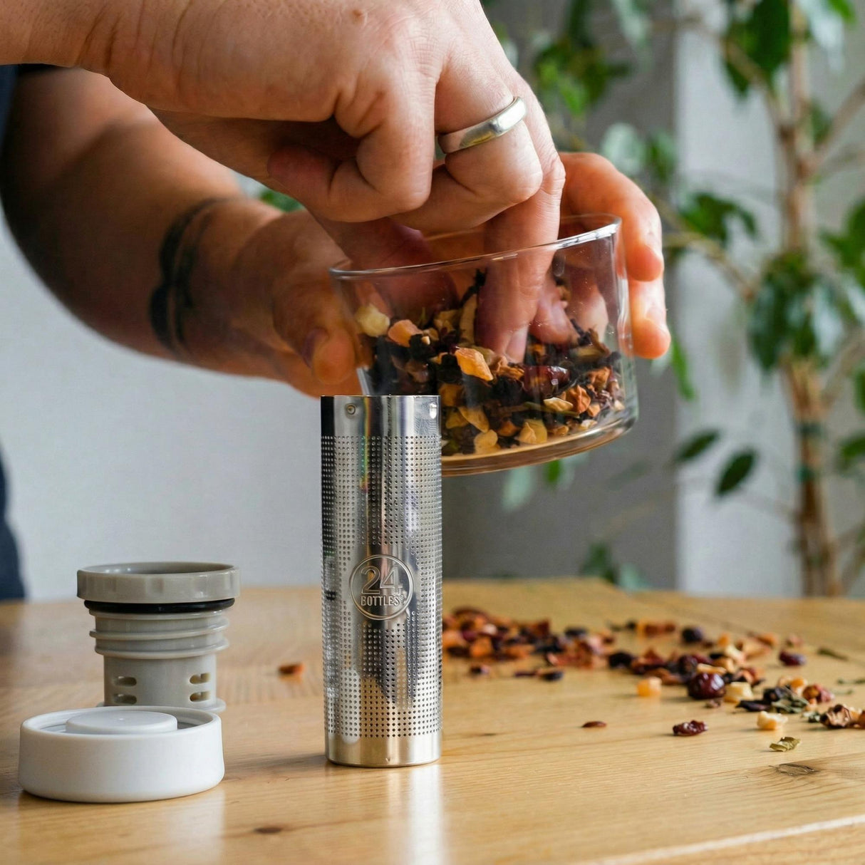 24Bottles Infuser lokk i grå og hvit plast og rustfritt stål, i bruk på et trebord med løs te