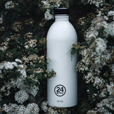 24Bottles Urban Drikkeflaske 1000 ml Ice White i rustfritt stål, stående blant grønne blader og hvite blomster