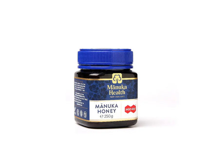 Et mørkt glass med blå skrukork og etikett i blått og gull, merket "Manuka Health New Zealand". Etiketten viser "MGO 400+" og teksten "Manuka Honey 250g", som indikerer høykvalitets manukahonning fra New Zealand.