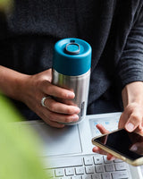 Black+Blum termokopp i rustfritt stål med blått lokk, holdt i hånden over laptop ved arbeidsbord.