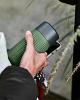 Black+Blum Glass Travel Cup 340 ml Olive i glass og silikon, holdt i hånden med telefon i bakgrunnen