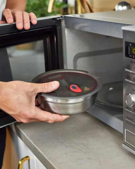 black+blum Matboks Rund 950ml Sølv i rustfritt stål, i bruk med lokket på vei inn i en mikrobølgeovn