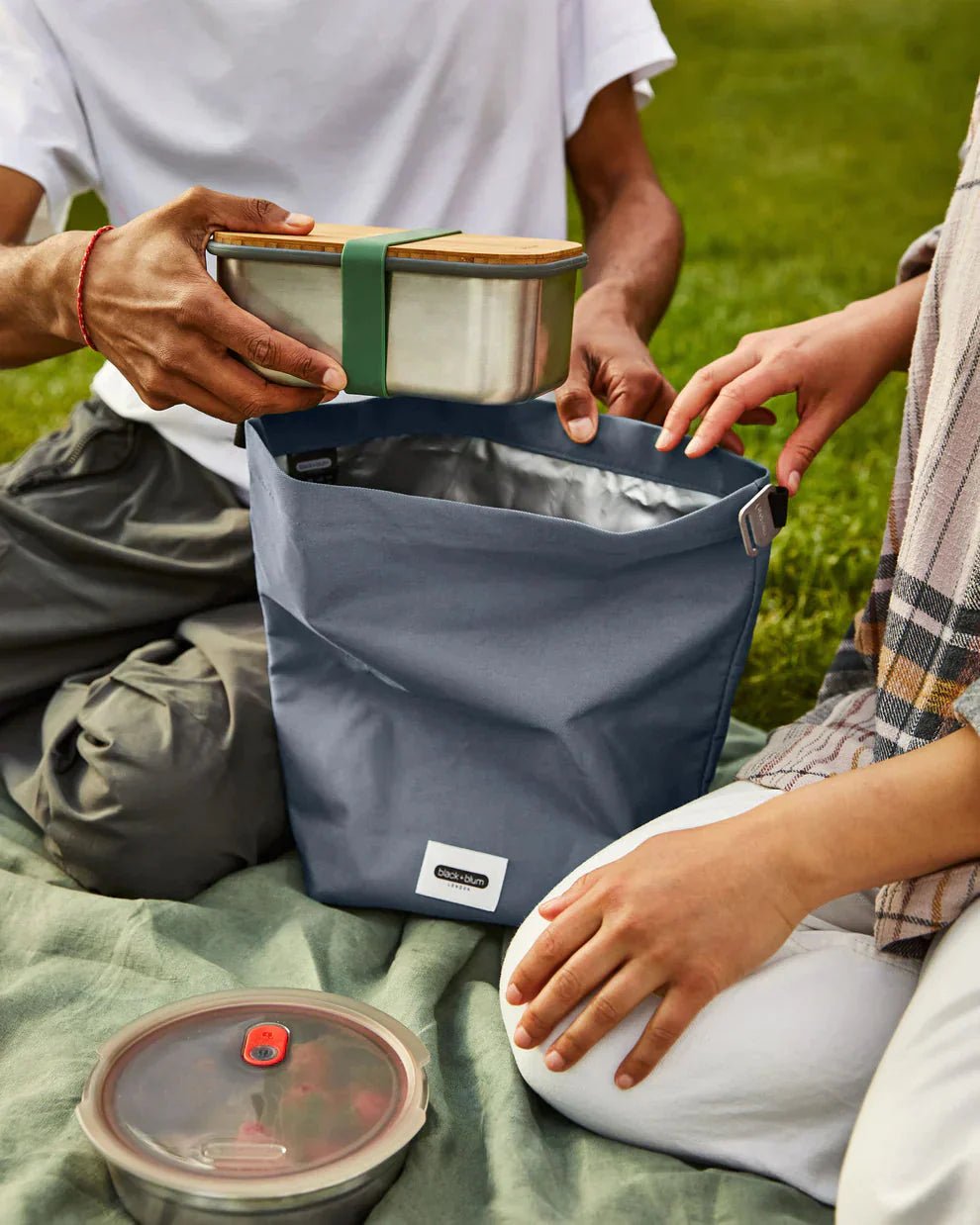 Black+Blum Matpose 6,7L Moss i resirkulert PET i bruk med matboks på et pledd i gresset