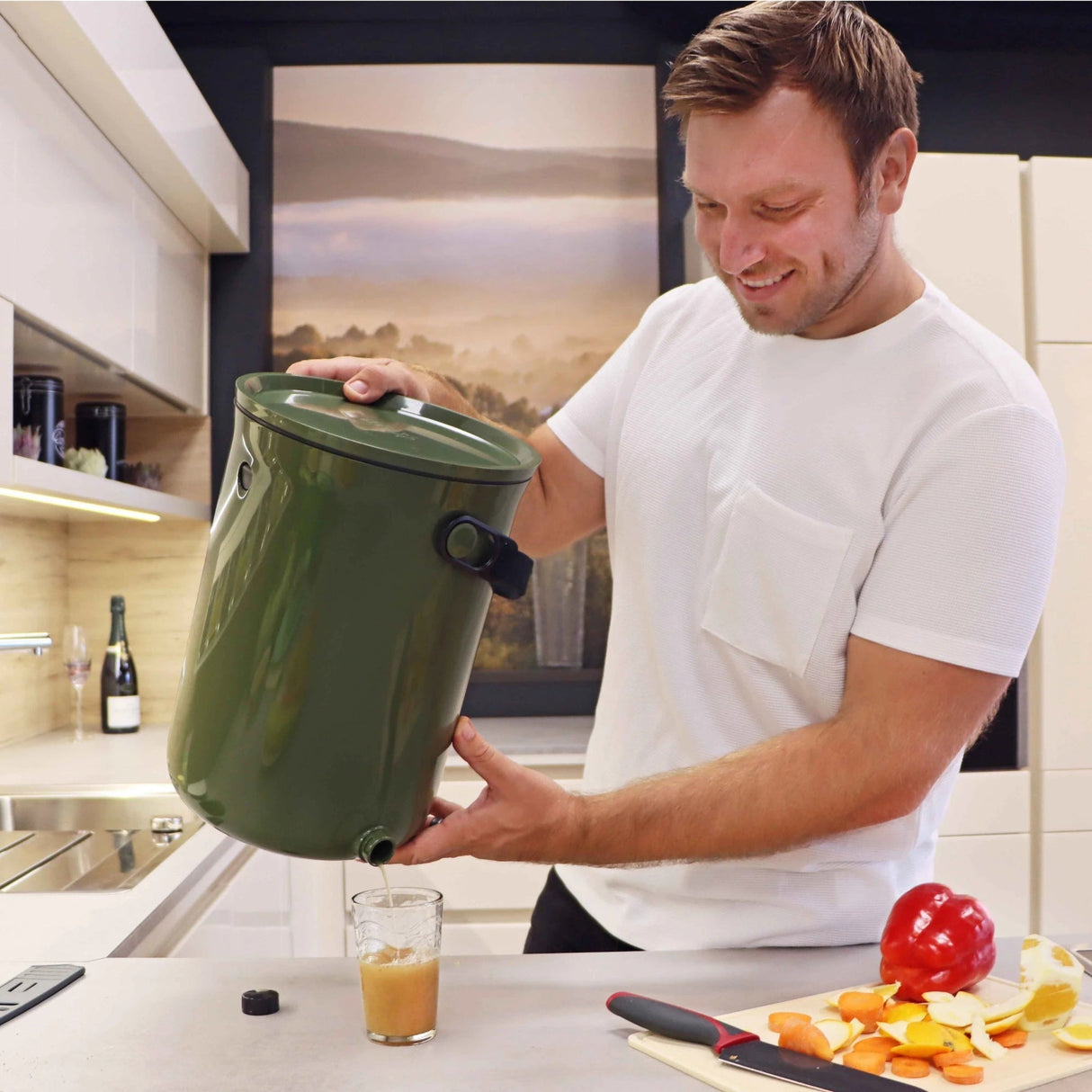 Bokashi Kompostbøtte 2.0 khaki i plast, en mann tapper væske fra bøtten ned i et glass på kjøkkenbenken