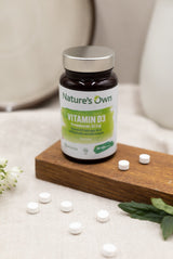Natures Own Vitamin D3 60 tabletter i brun flaske med grønn etikett på treplate med blomster og tabletter