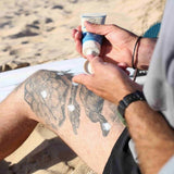 Suntribe Mineral Solkrem SPF 50, 100 ml, beige og blå i tube, i bruk på en tatovert legg på stranden