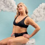 Wuka Stretch Cotton Midi Brief Heavy Flow i svart bomull, en kvinne sitter i skyer med armen i siden